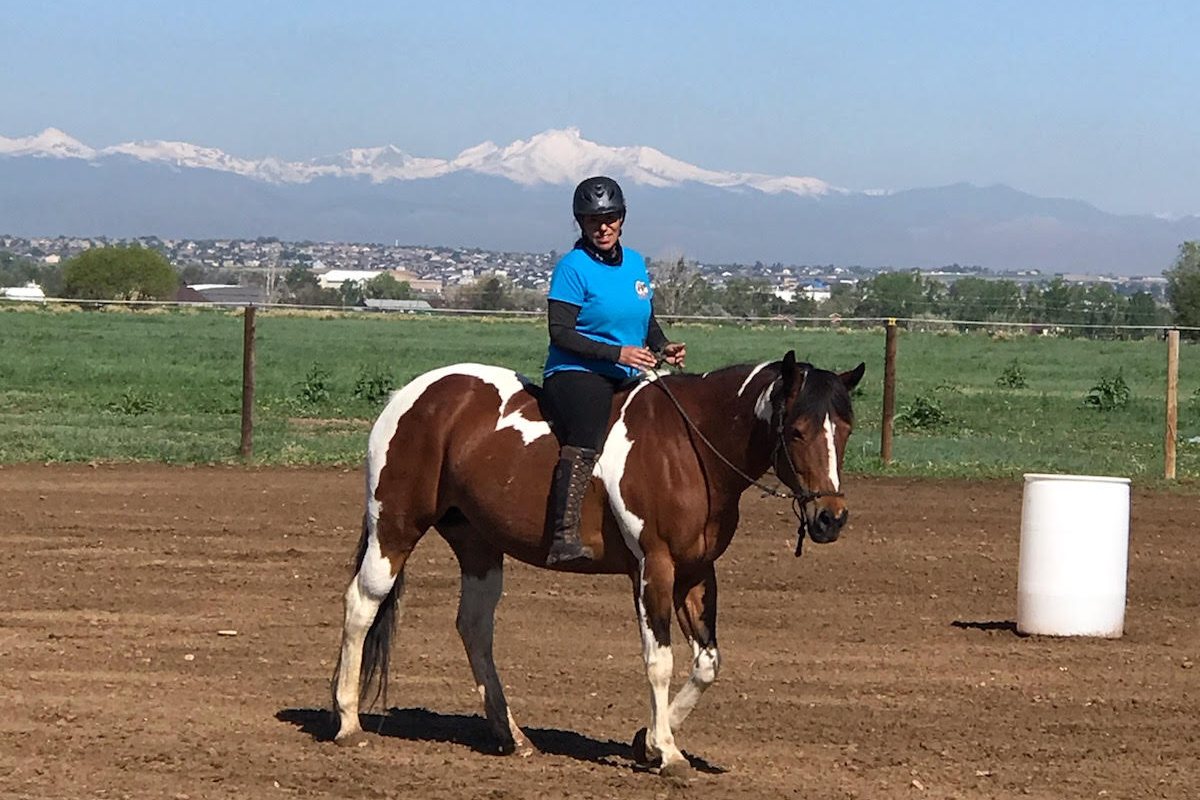 Carla riding Bourbon, her brown and white Paint mare, in the arena at SonFlower Ranch with the snowy Rocky Mountains behind them