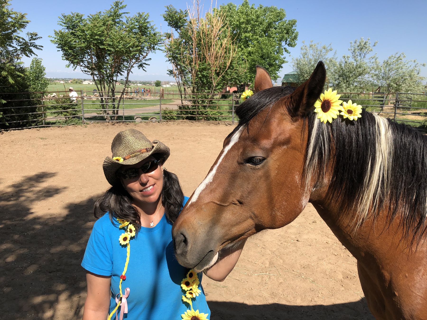 Carla smiling beside Bourbon, a brown and white Paint horse wearing sunflowers in her mane