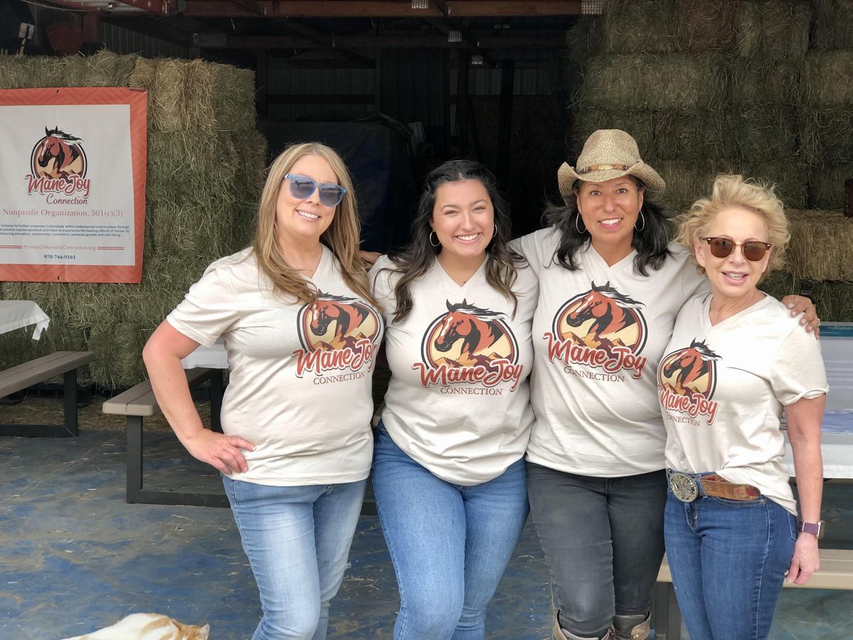 Four smiling women wearing Mane Joy Connection T-shirts in a hay barn
